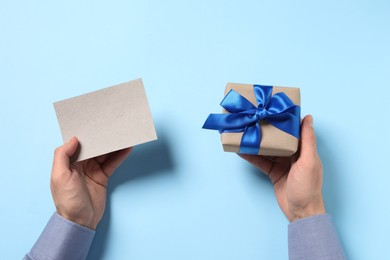 Photo of Man holding gift box with bow and blank greeting card on light blue background, top view