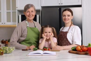 Photo of Cute little girl with her mother and grandmother cooking by recipe book in kitchen