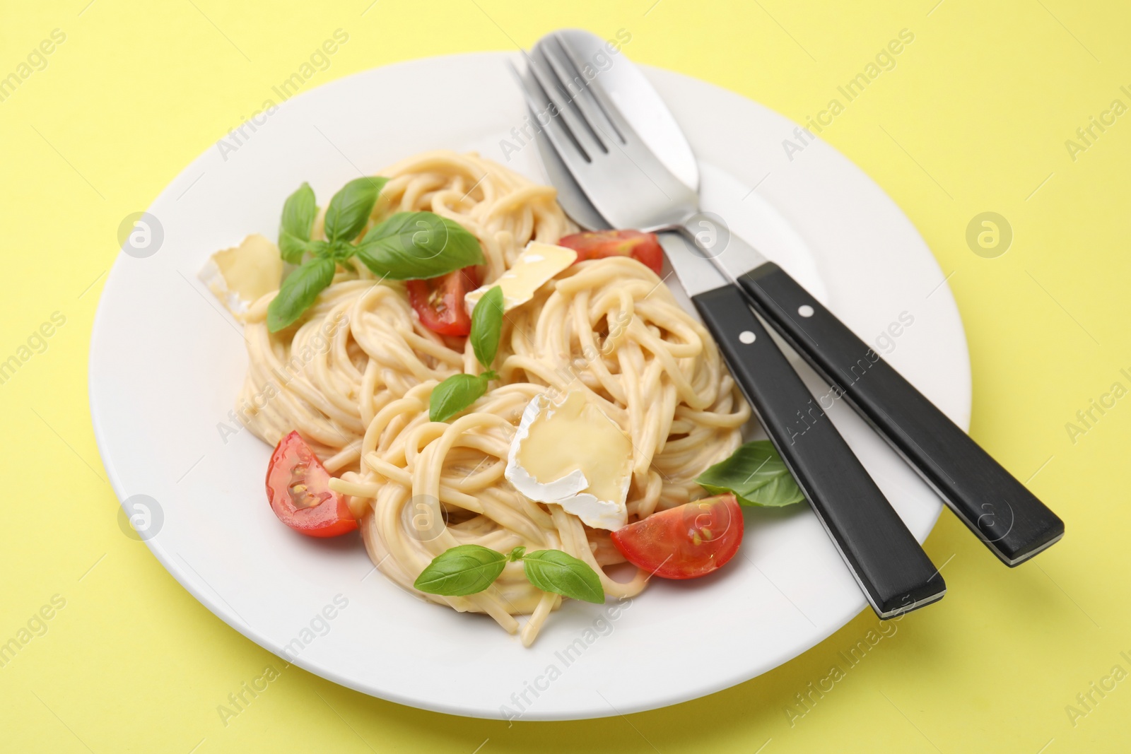 Photo of Delicious pasta with brie cheese, tomatoes, basil and cutlery on yellow table, closeup