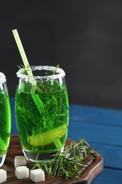 Photo of Glass of homemade refreshing tarragon drink, sprigs and sugar cubes on blue wooden table