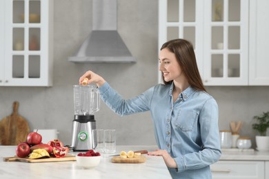 Beautiful young woman adding banana into blender for tasty smoothie in kitchen