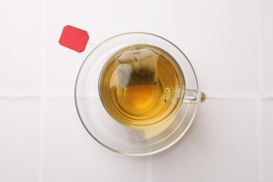 Tea bag in cup with hot drink on white tiled table, top view