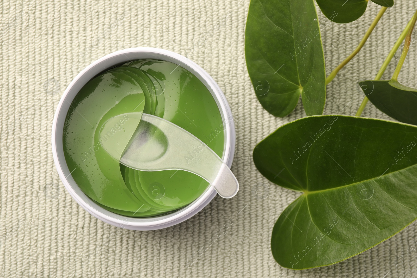 Photo of Jar of under eye patches with spoon and green leaves on light cloth, flat lay. Cosmetic product