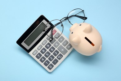 Photo of Calculator, glasses and piggy bank on light blue background, flat lay