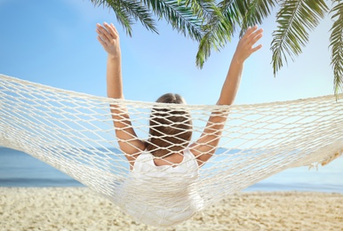Image of Woman relaxing in hammock under green palm leaves on sunlit beach
