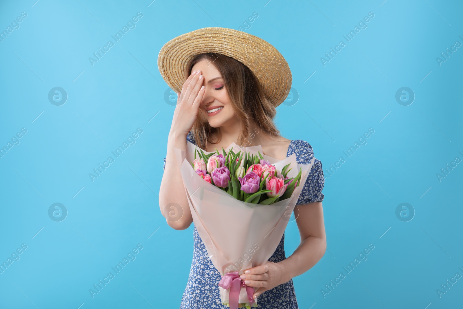 Photo of Happy young woman in straw hat holding bouquet of beautiful tulips on light blue background