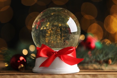 Photo of Beautiful snow globe on wooden table against blurred Christmas lights, closeup