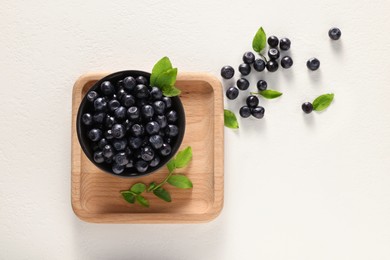 Tasty fresh bilberries with green leaves and bowl on white table, flat lay