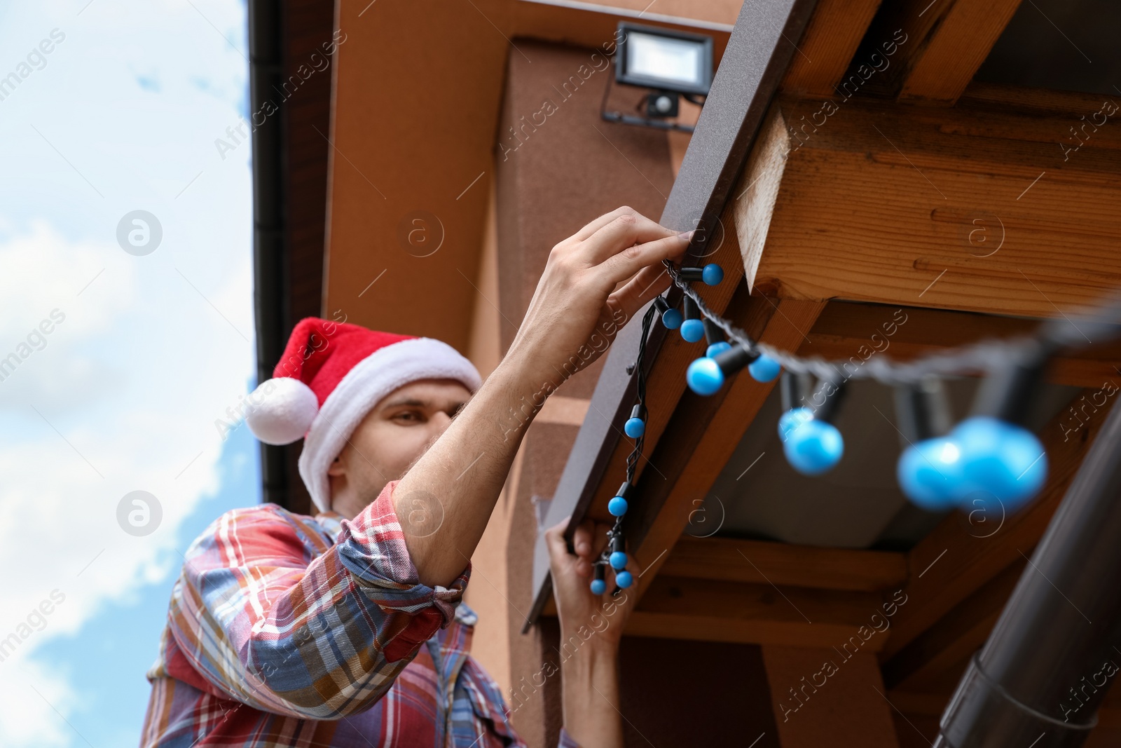 Photo of Man in Santa hat decorating house with Christmas lights outdoors, low angle view