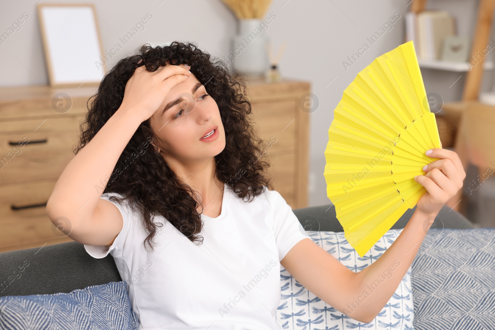 Photo of Young woman waving yellow hand fan to cool herself on sofa at home