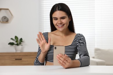 Happy young woman having video chat via smartphone and waving hello at table in room