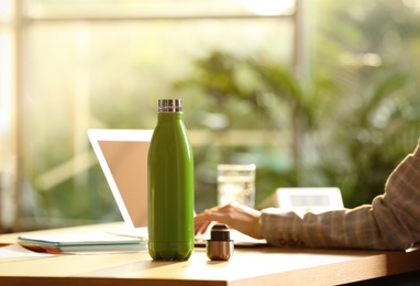 Photo of Woman working at table in modern office, focus on green thermos bottle