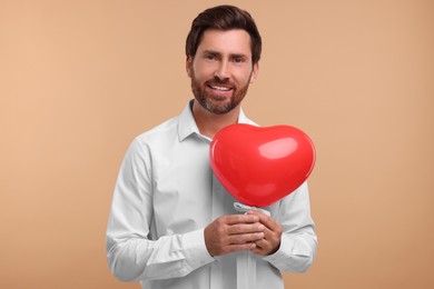 Handsome bearded man holding red heart shaped balloon on beige background