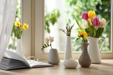 Different beautiful spring flowers, burning candle and book on window sill
