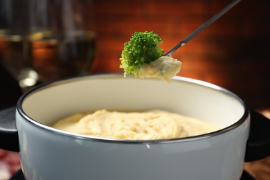 Dipping piece of broccoli into fondue pot with melted cheese on blurred background, closeup