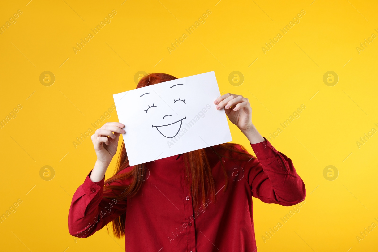 Photo of Woman hiding behind sheet of paper with happy face on yellow background