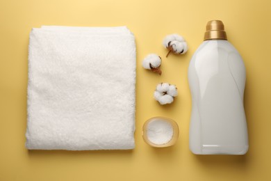 Photo of Bottle of fabric softener, terry towel, detergent powder and cotton flowers on pale yellow background, flat lay