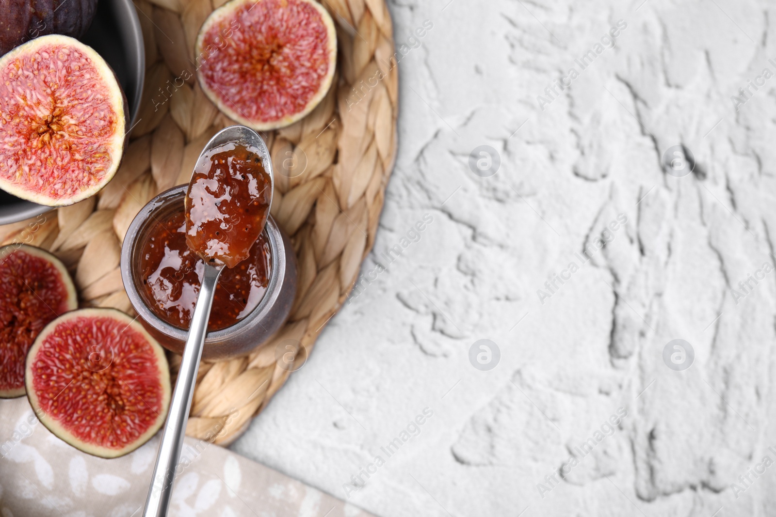 Photo of Glass jar of tasty sweet fig jam with spoon and fruits on white textured table, flat lay. Space for text