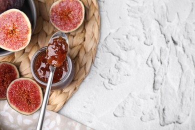 Glass jar of tasty sweet fig jam with spoon and fruits on white textured table, flat lay. Space for text