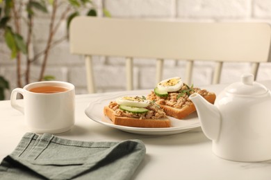 Photo of Delicious sandwiches with tuna, vegetables, boiled egg and cup of tea on white table indoors. Tasty breakfast