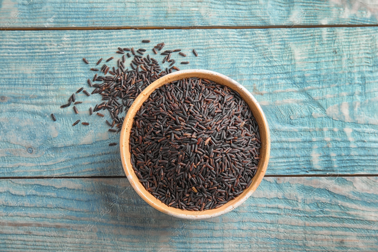 Photo of Bowl with uncooked black rice on blue wooden background, top view