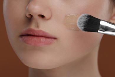 Photo of Teenage girl applying foundation on face with brush against brown background, closeup