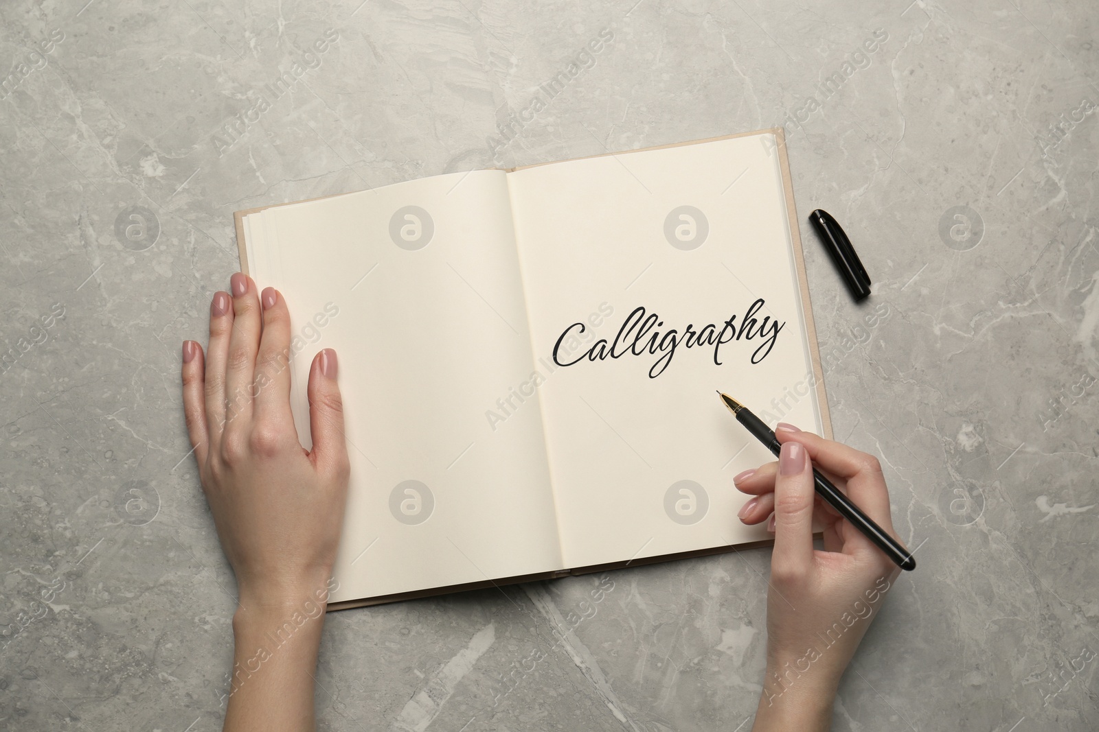 Image of Woman writing word Calligraphy in notebook at grey marble table, top view