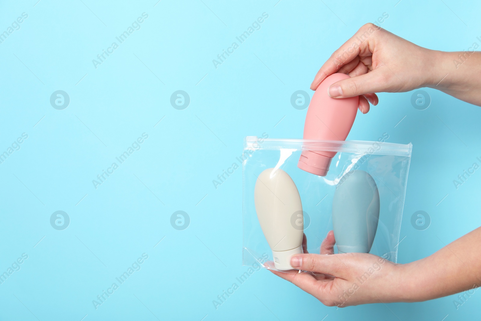 Photo of Cosmetic travel kit. Woman putting small bottle with personal care product into plastic bag against light blue background, closeup. Space for text