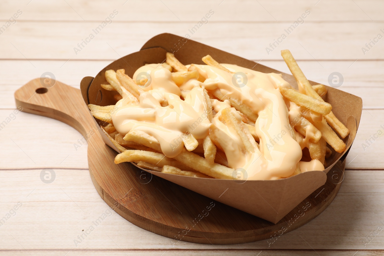 Photo of Tasty potato fries and cheese sauce in paper container on light wooden table, closeup