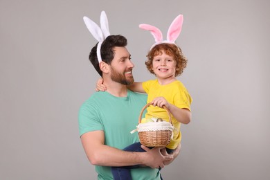 Photo of Happy father and son wearing cute bunny ears headbands on light grey background. Boy holding Easter basket with painted eggs