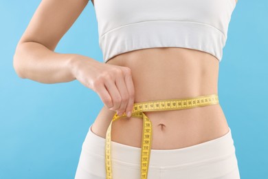 Photo of Slim woman measuring waist with tape on light blue background, closeup. Weight loss