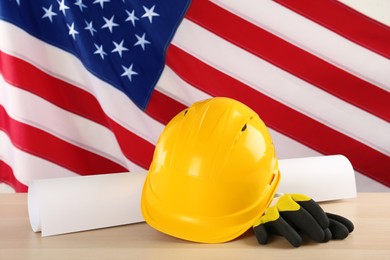 Photo of Yellow hard hat, gloves and paper on light wooden table against American flag