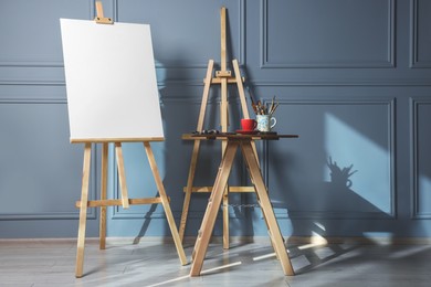 Photo of Easel with blank canvas and different art supplies on wooden table near grey wall indoors