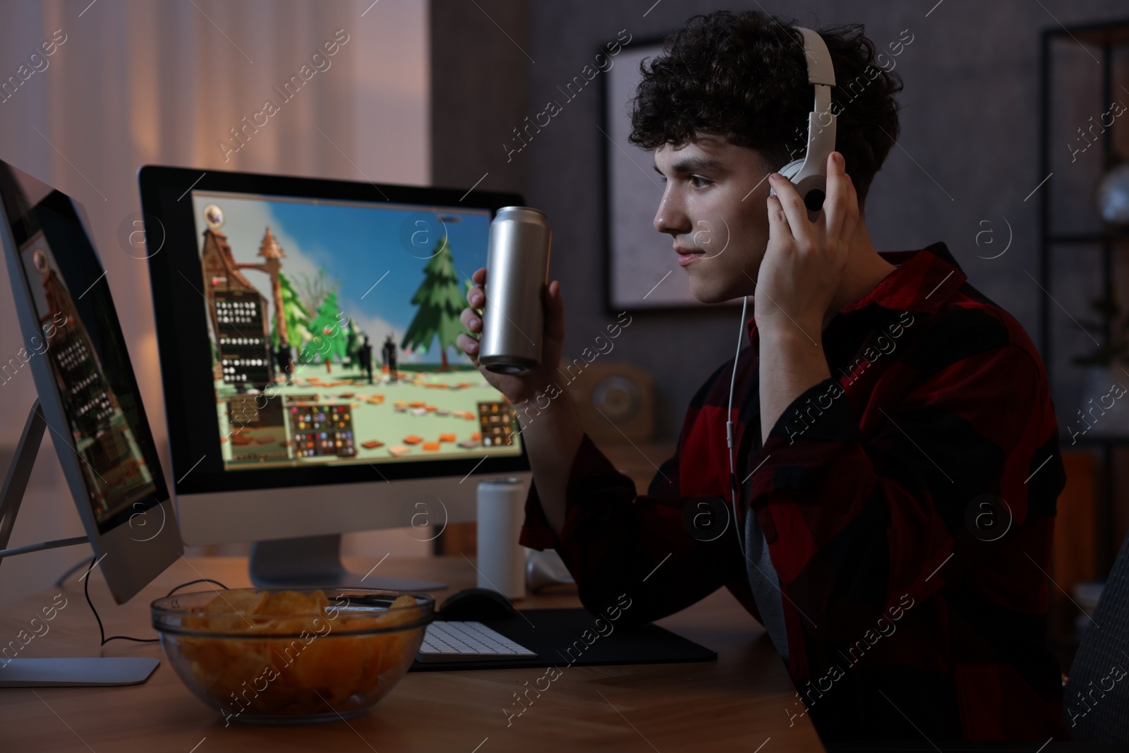 Photo of Young man with energy drink and headphones playing video game at wooden desk indoors