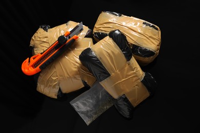 Smuggling, drug trafficking. Packages with narcotics and utility knife on black surface, flat lay