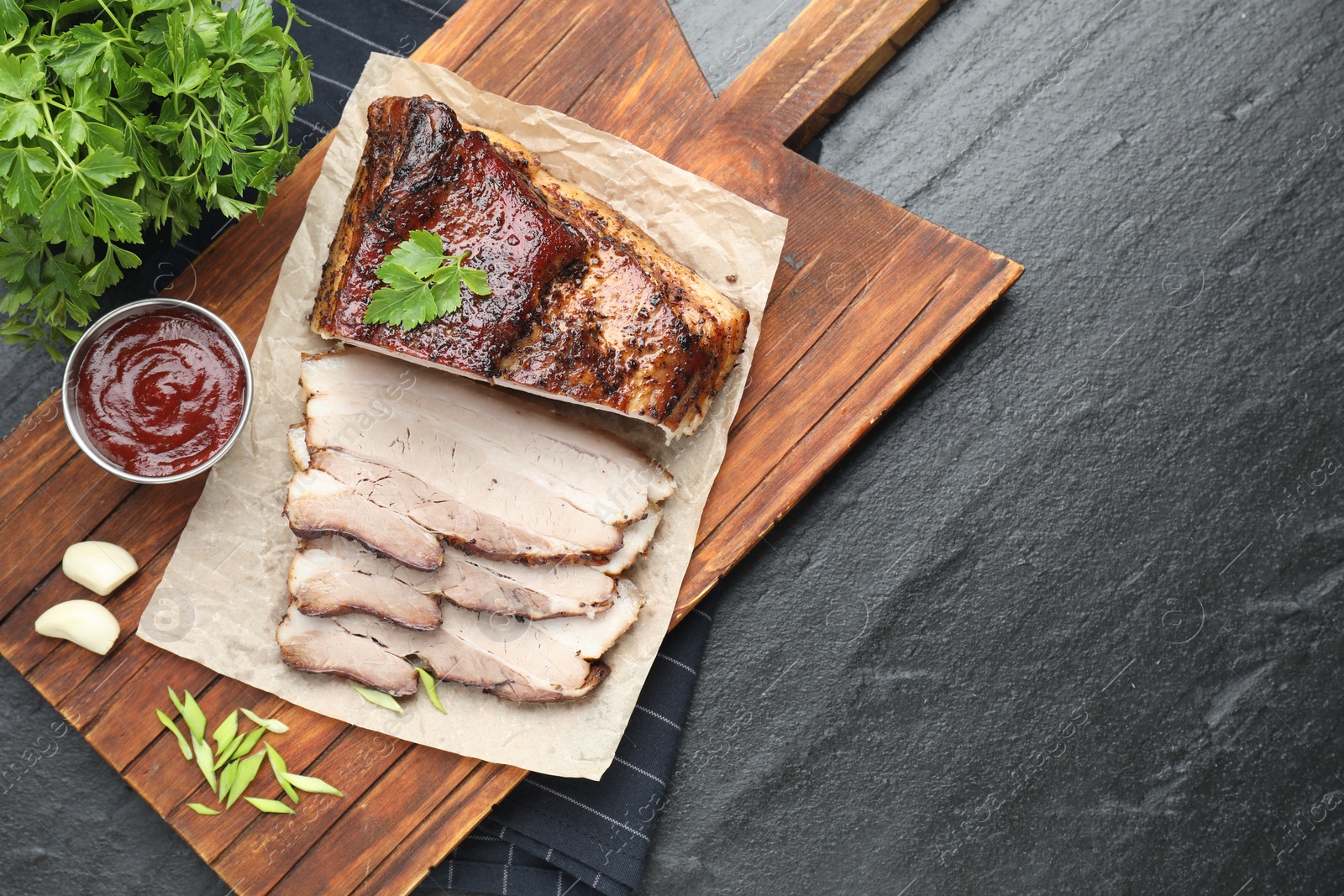 Photo of Pieces of baked pork belly served with sauce and parsley on black textured table, top view. Space for text