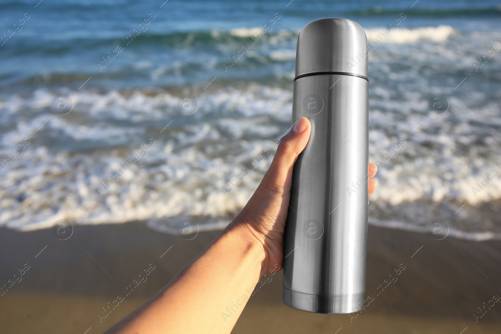 Photo of Woman holding metallic thermos with hot drink on beach near sea, closeup