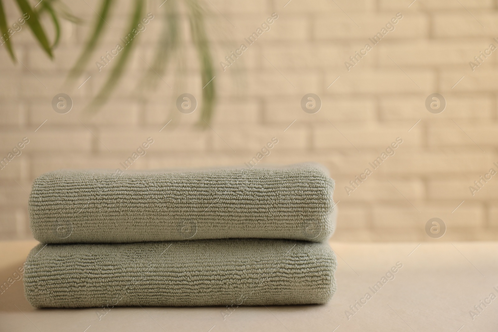 Photo of Stacked soft towels and green leaves on white table near brick wall indoors, space for text