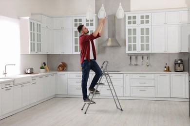 Photo of Young man installing ceiling lamp on stepladder in kitchen