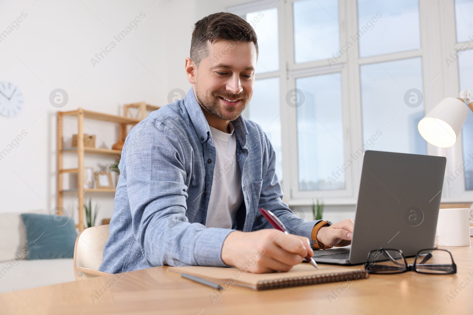 Photo of Happy man writing notes while working on laptop at wooden desk in room