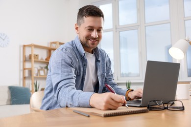 Happy man writing notes while working on laptop at wooden desk in room