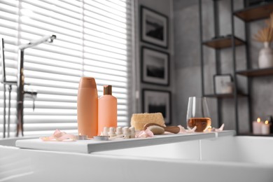 Photo of Wooden tray with wine, toiletries and flower petals on bathtub in bathroom