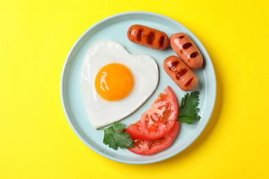 Plate of heart shaped fried egg and sausages on yellow background, top view