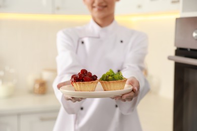 Photo of Happy professional confectioner holding plate with delicious tartlets in kitchen, closeup