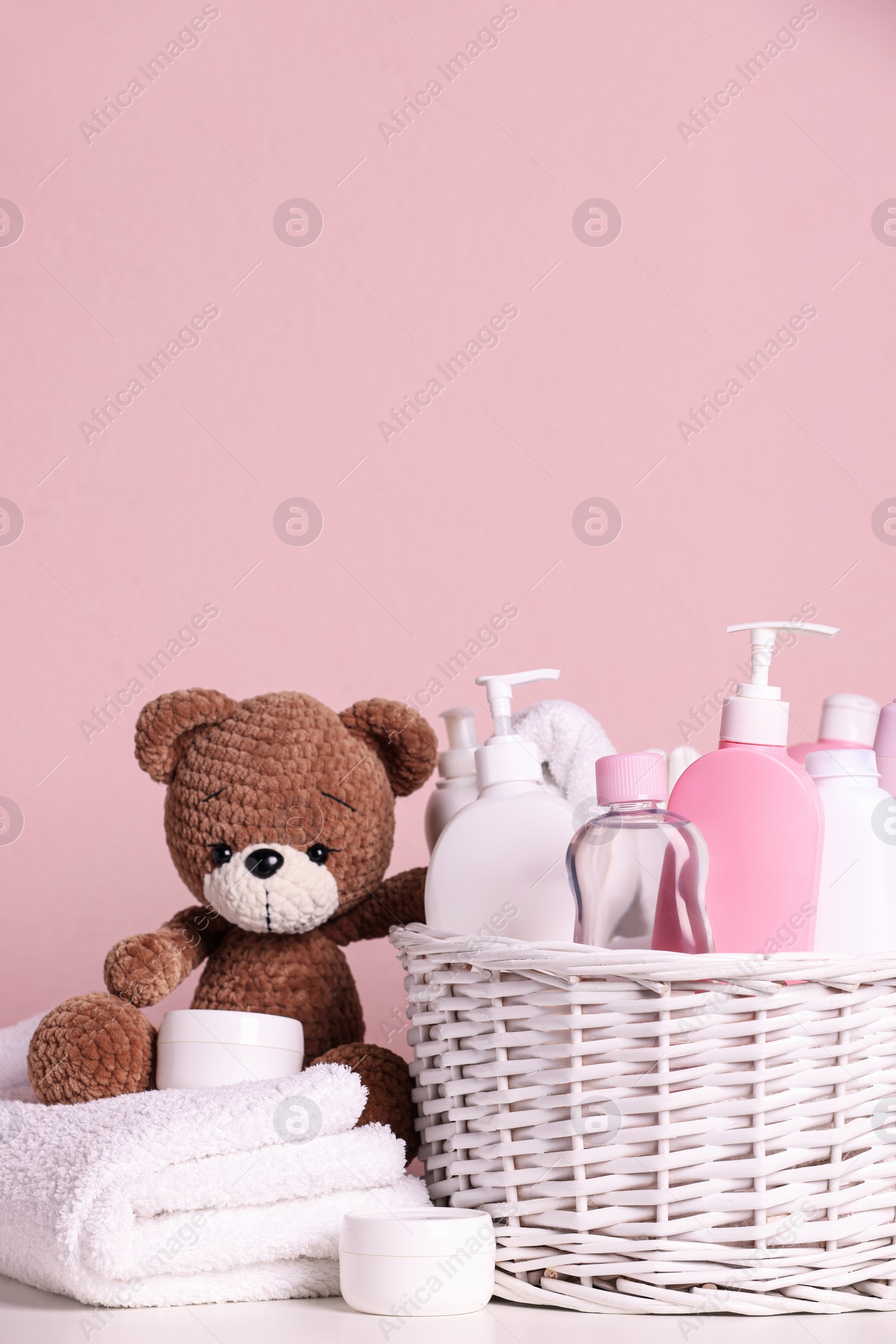 Photo of Baby cosmetic products in wicker basket, bath accessories and knitted toy bear on white table against pink background