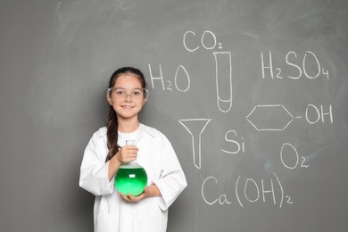 Photo of Little school child in laboratory uniform with flask of liquid and chemical formulas on grey background