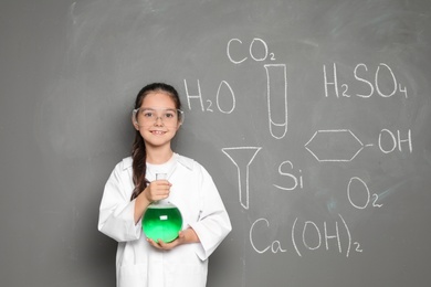 Little school child in laboratory uniform with flask of liquid and chemical formulas on grey background