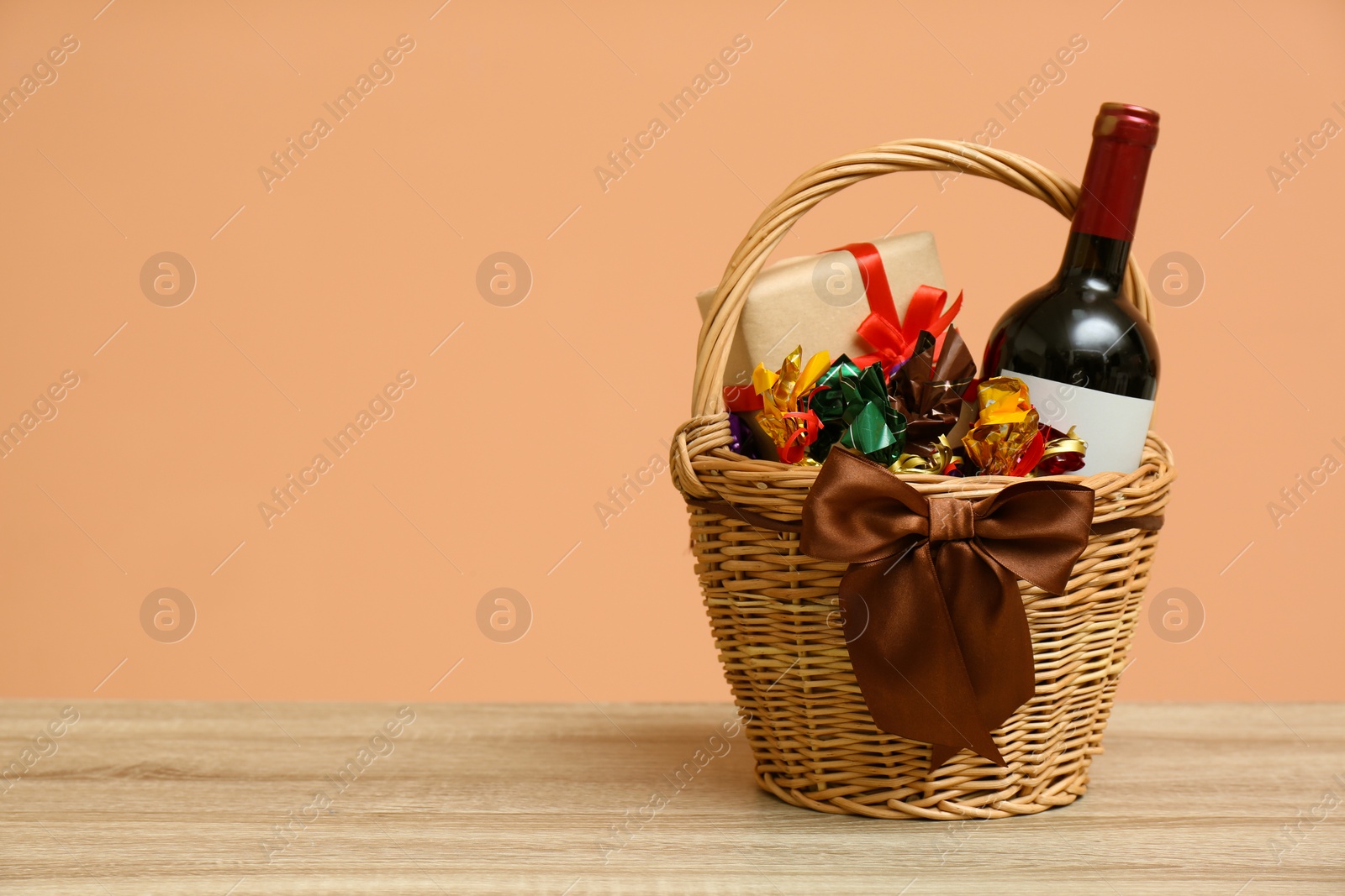 Photo of Wicker gift basket with food and wine on wooden table. Space for text