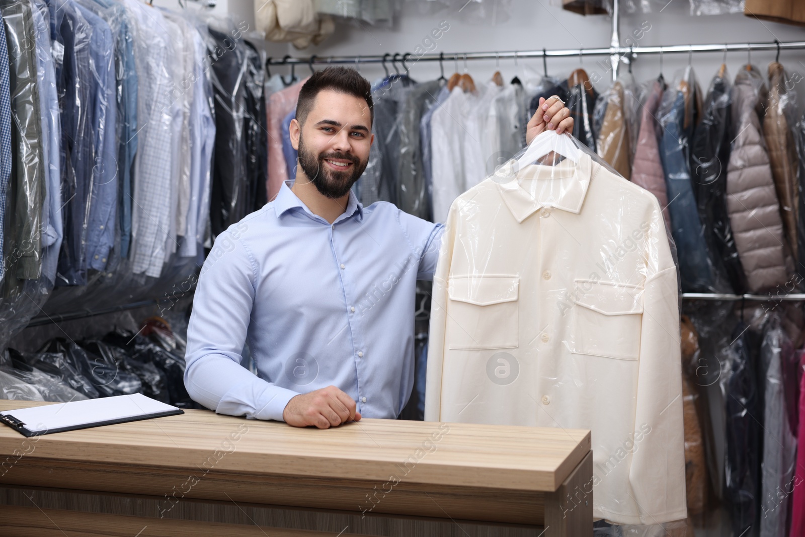 Photo of Dry-cleaning service. Happy worker holding hanger with shirt in plastic bag at counter indoors