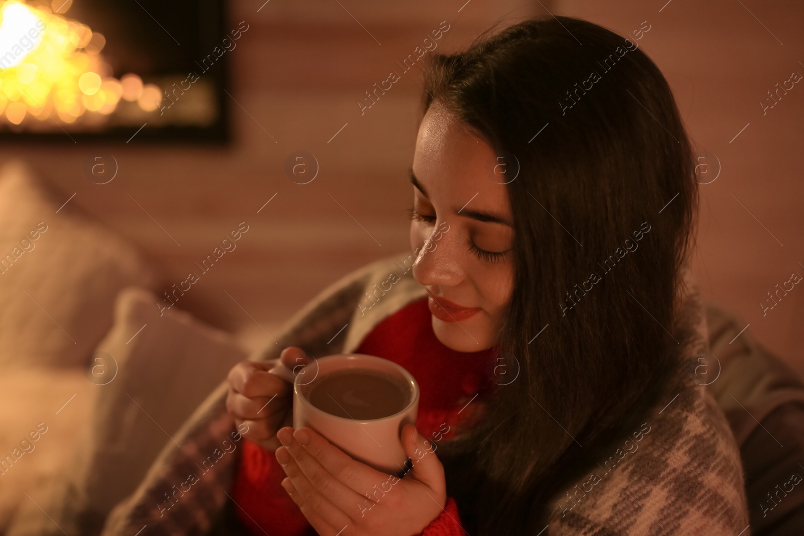 Photo of Young woman drinking coffee near decorative fireplace at home. Winter season